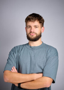 Portrait of a man with short brown hair and a beard, wearing a blue-gray T-shirt, arms crossed against a neutral gray studio background.